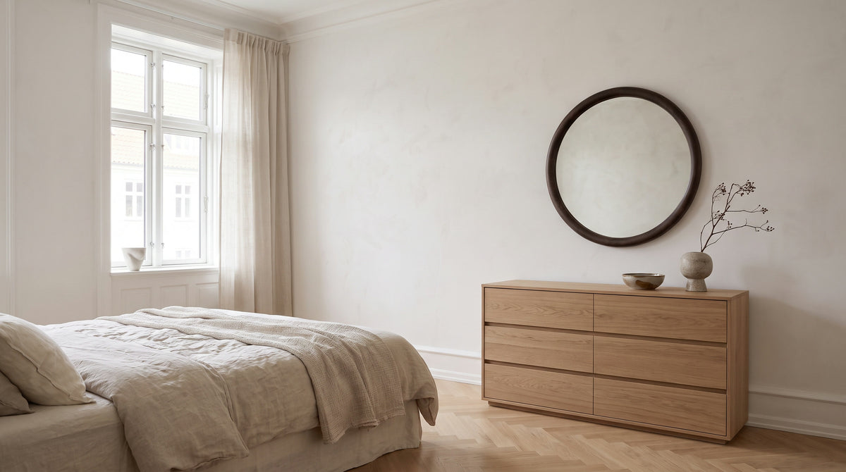Serene bedroom with linen bedding and a round walnut-framed mirror above an oak chest of drawers