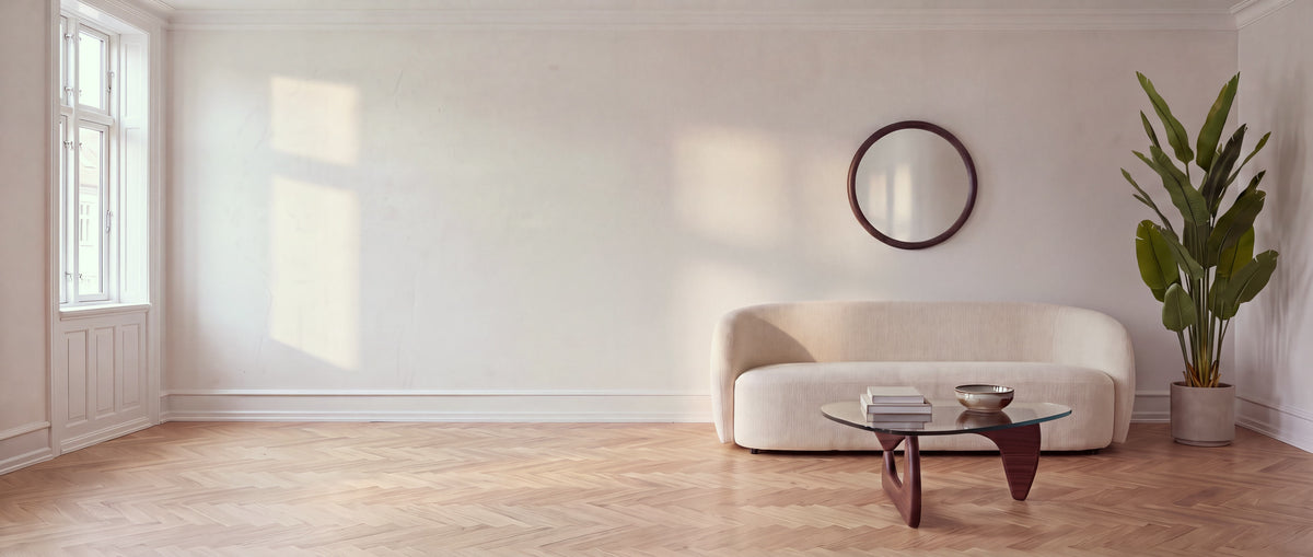 Calm, light-filled living room with a walnut and glass coffee table, a round walnut-framed mirror above a cream sofa and a tall bird-of-paradise plant